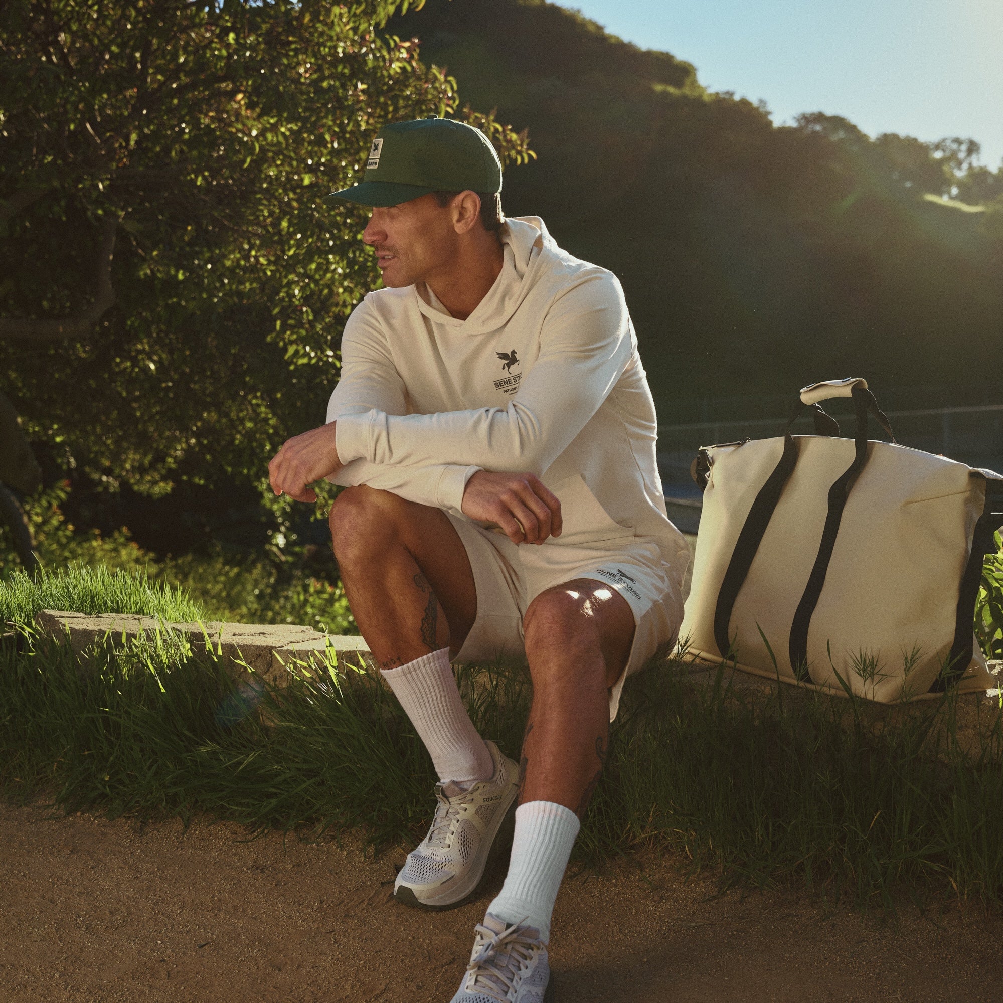 Man sitting outdoors with a white bag next to him, wearing a green cap and white outfit Sene hat model Tyler in Court Green