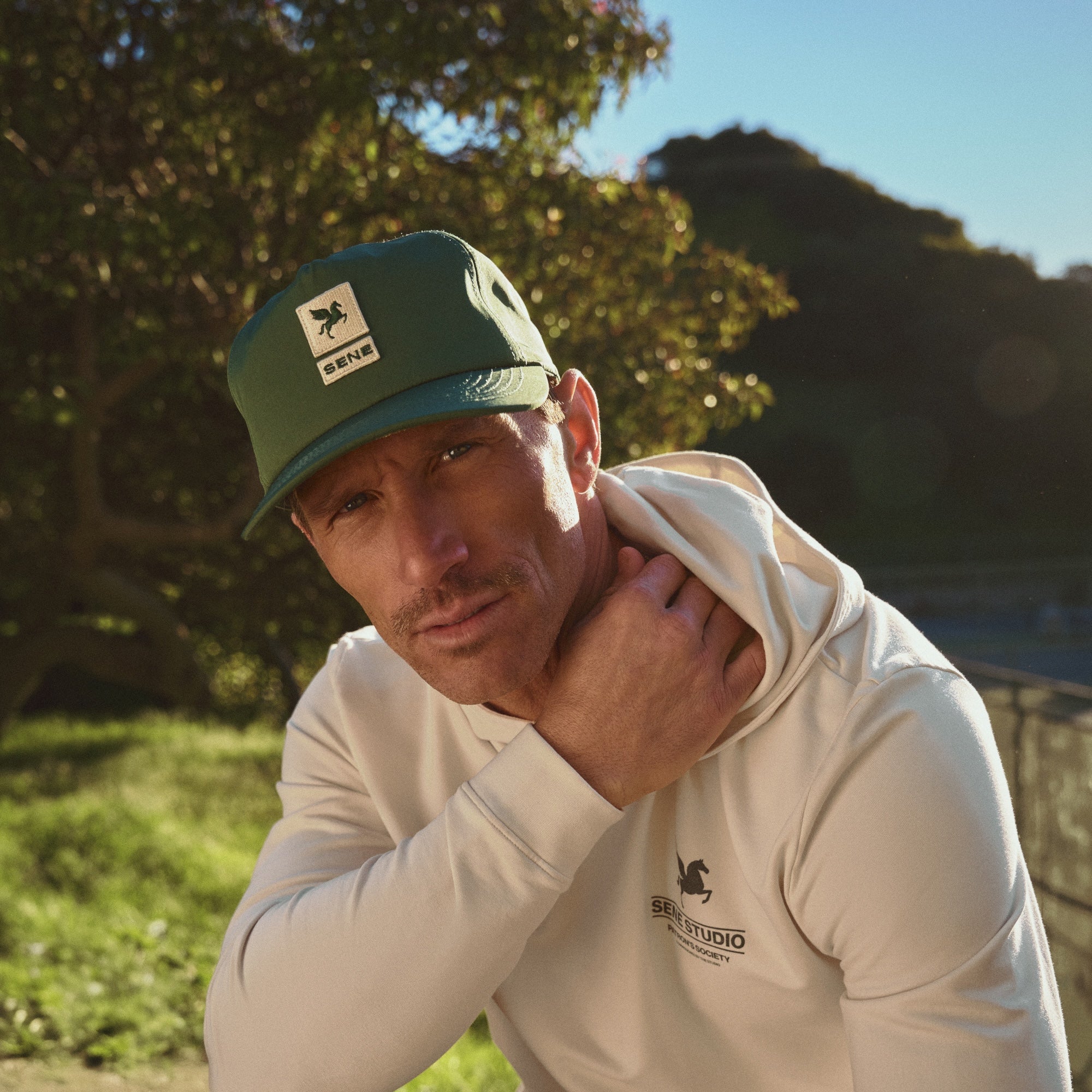 Man wearing a green cap and white hoodie with logos, sitting outdoors Sene hat model Tyler in Court Green