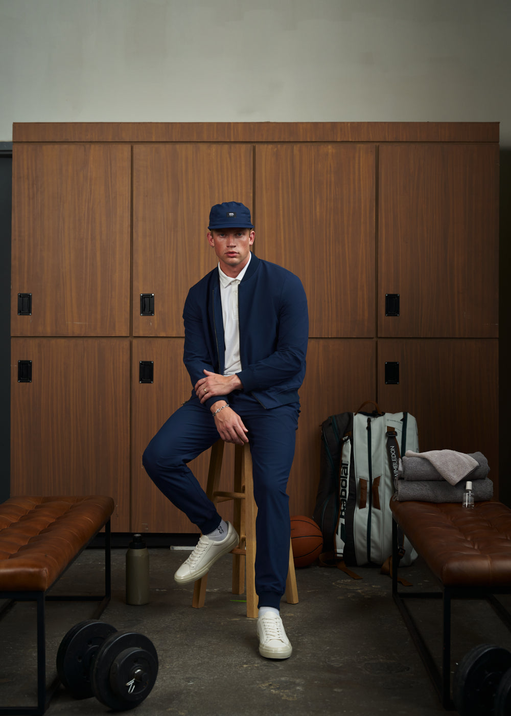 Man in blue athletic wear sitting in front of wooden lockers with gym equipment around him.