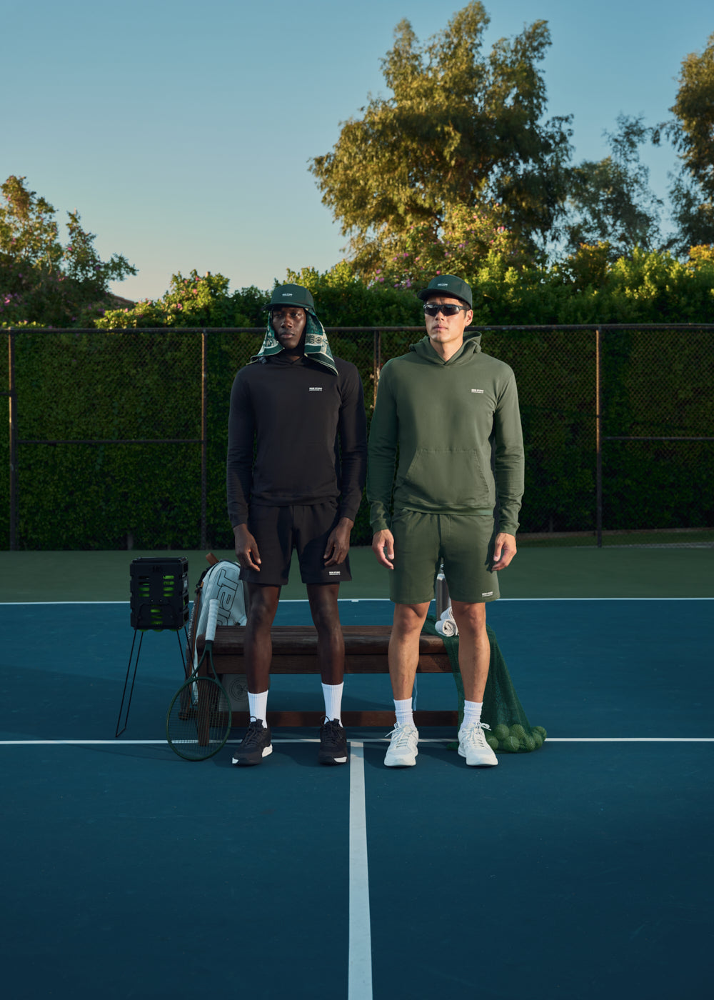 Two men standing on a tennis court, dressed in athletic wear with equipment nearby.