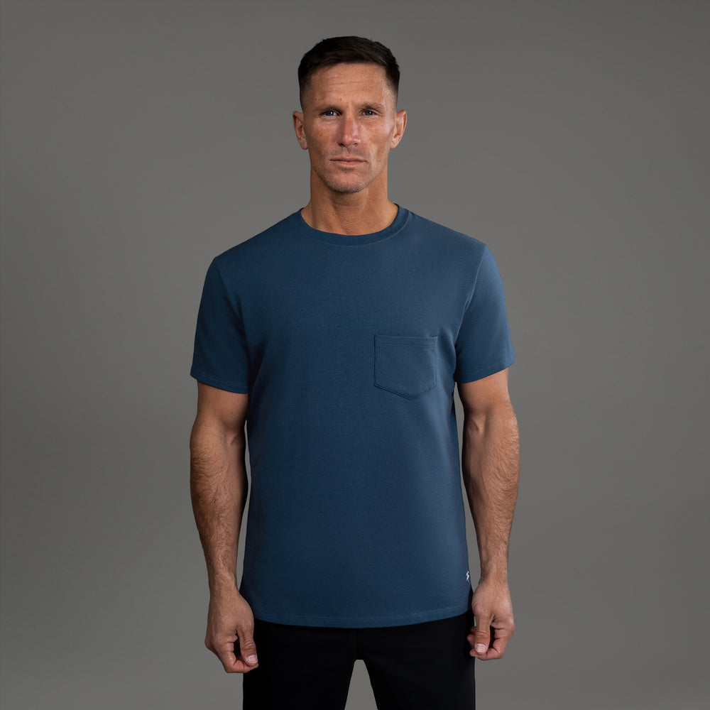 Man wearing a blue T-shirt with a pocket, standing against a gray background.