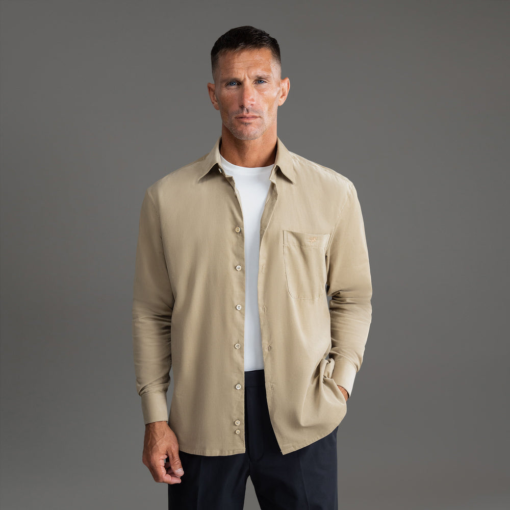 A man in a beige button-up shirt and dark pants stands against a gray background.