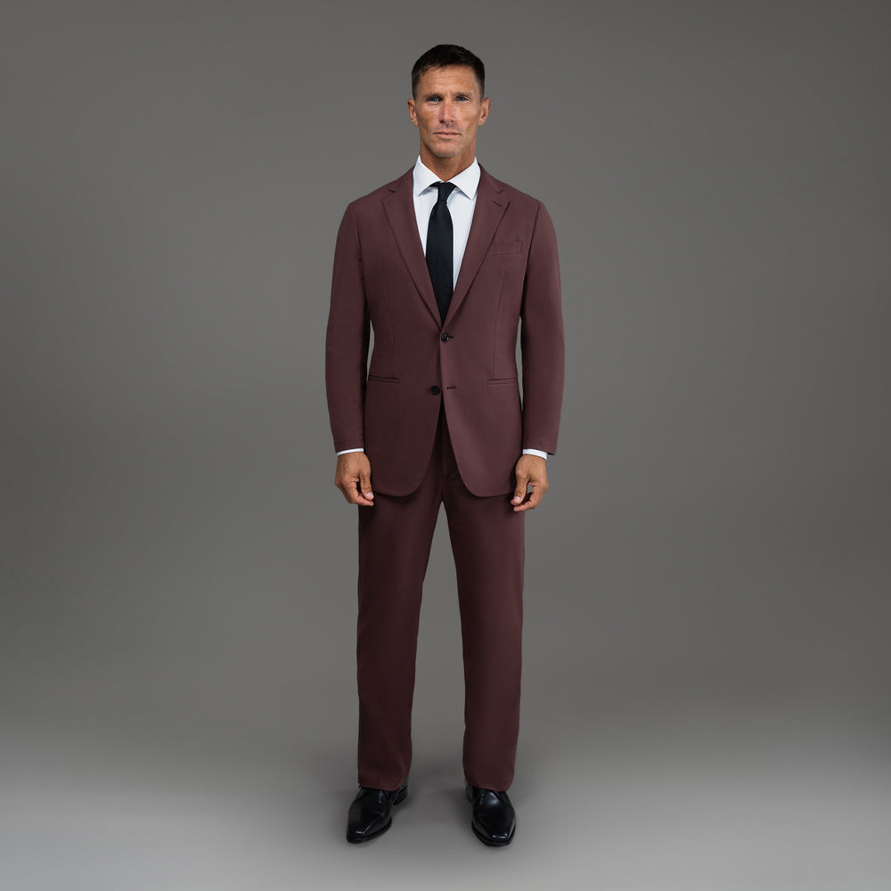 Man in a burgundy suit with a black tie standing against a neutral background.