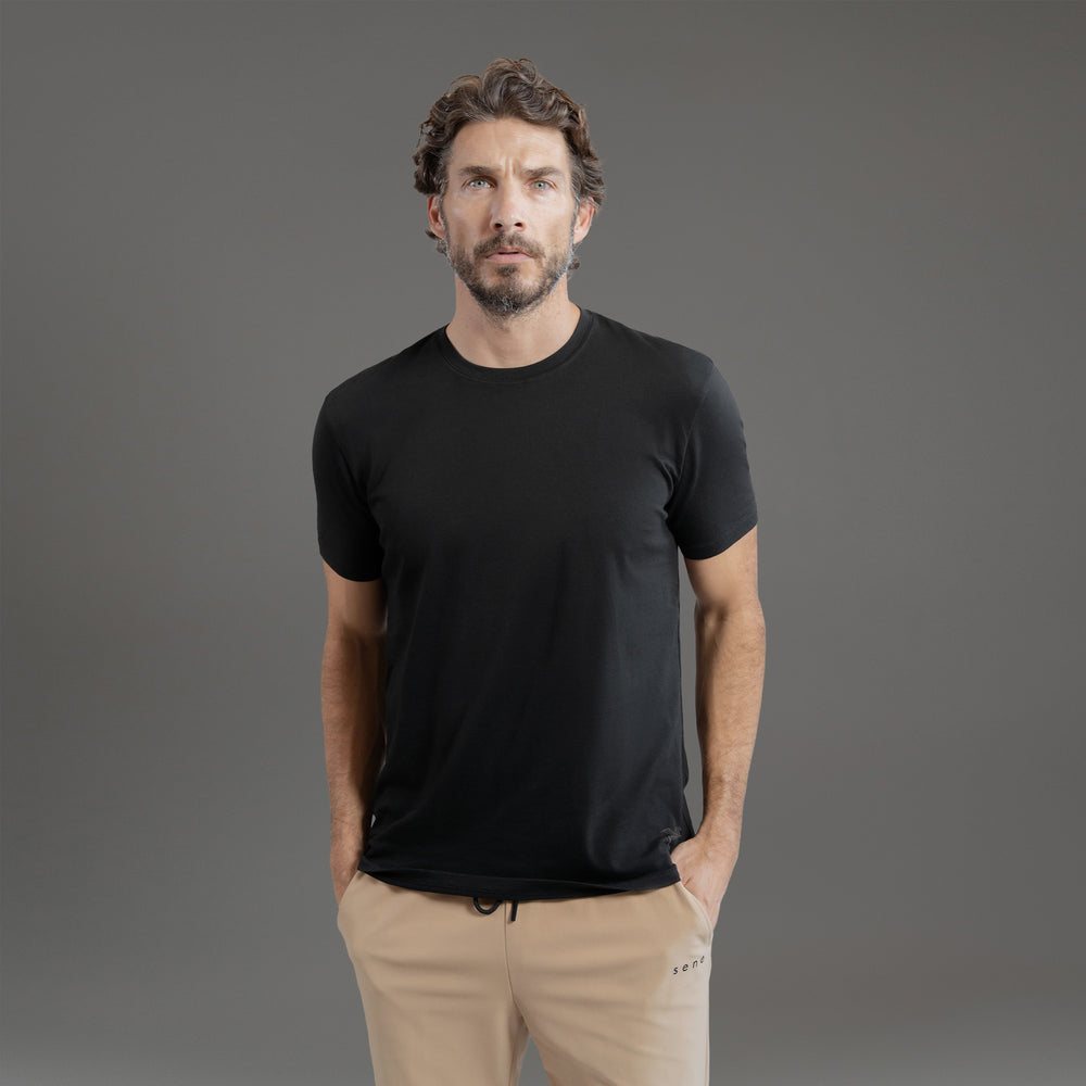 Man in black t-shirt and beige pants standing against a gray background.