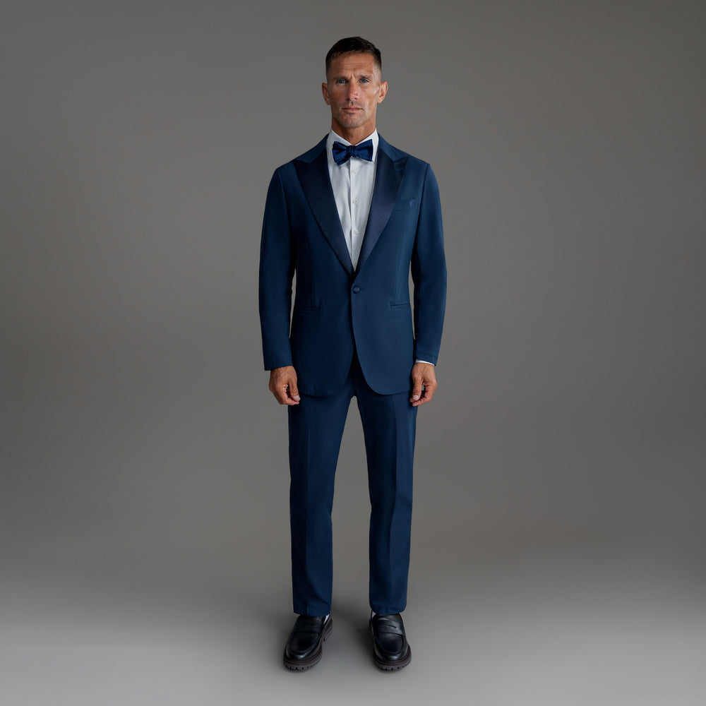 Man in a blue suit with a bow tie standing against a gray background.