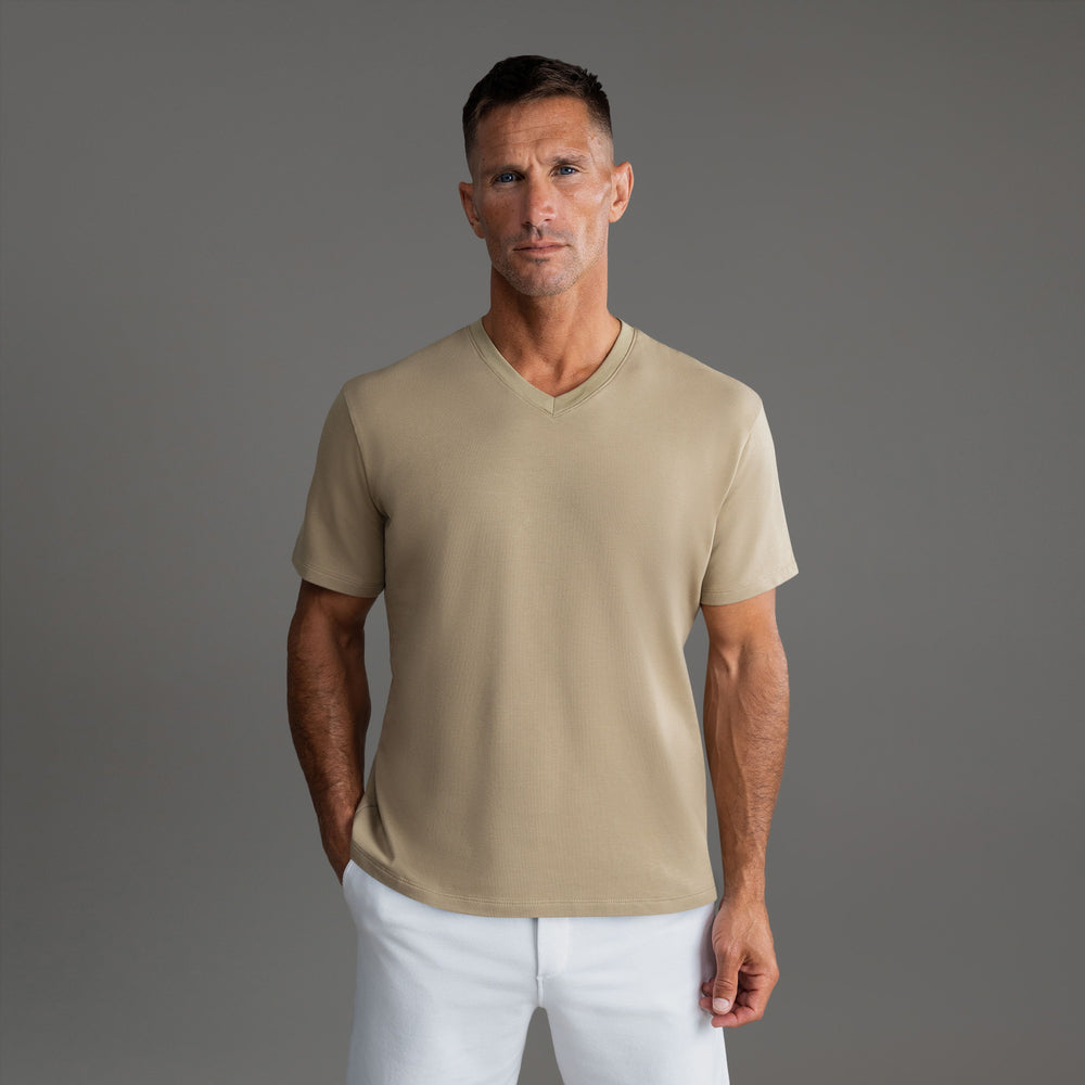 Man in a tan V-neck t-shirt and white pants against a gray background.