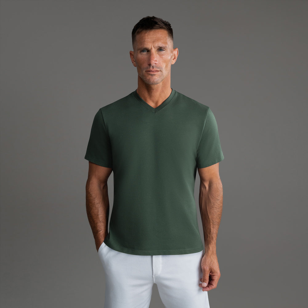 Man in a green V-neck shirt and white pants posing against a grey background.