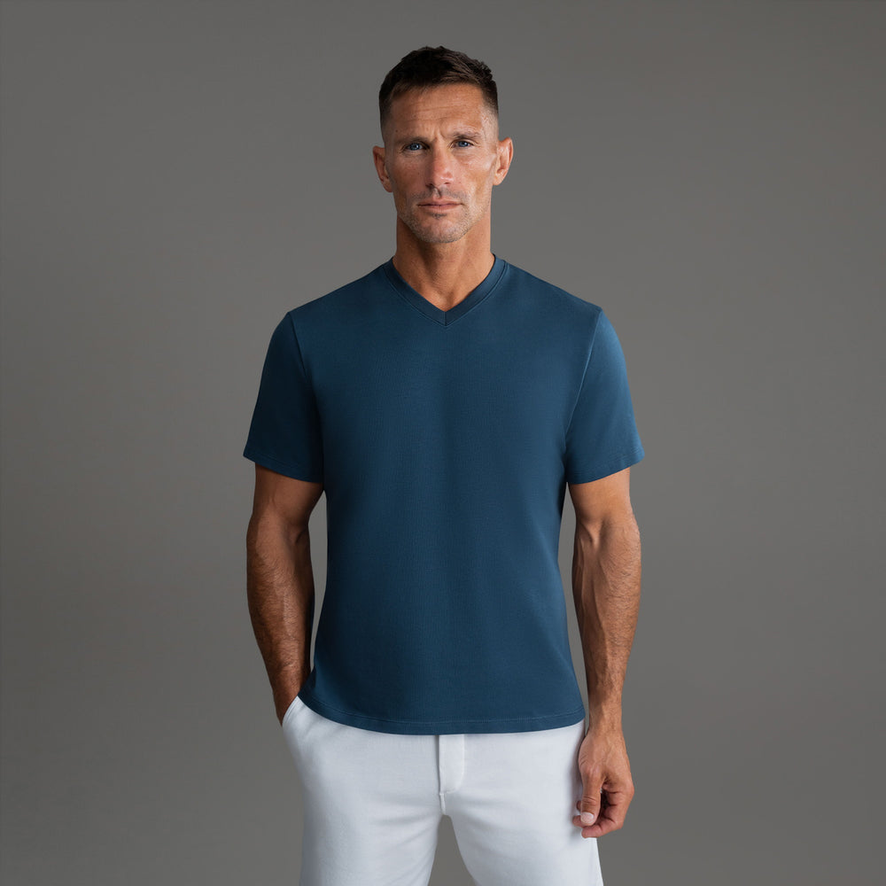 Man wearing a blue V-neck t-shirt and white pants against a neutral background.