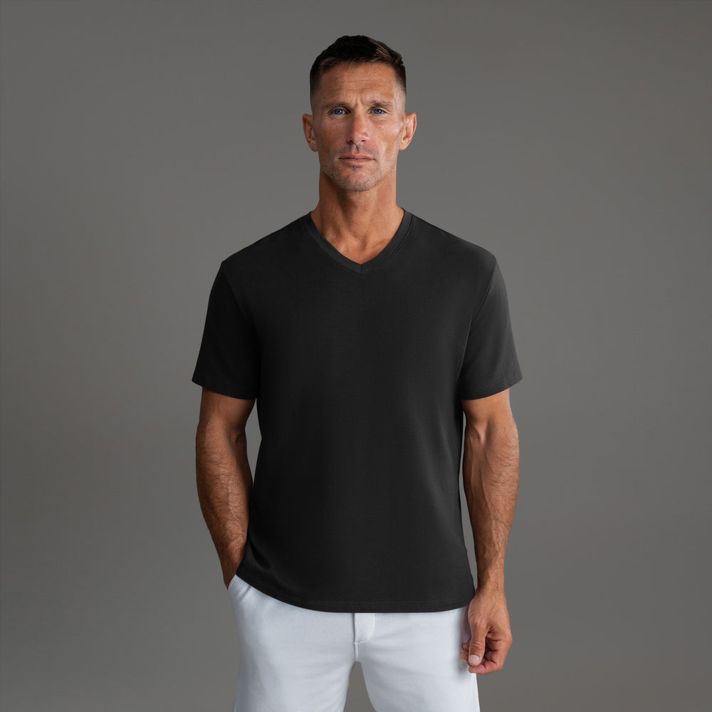 Man in a black V-neck T-shirt and white pants, standing against a gray background.