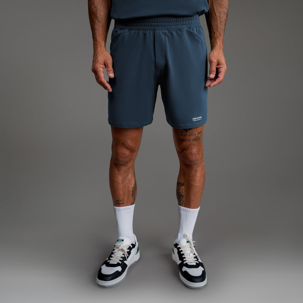 Man wearing blue shorts, white socks, and sneakers, tattoos visible on legs.