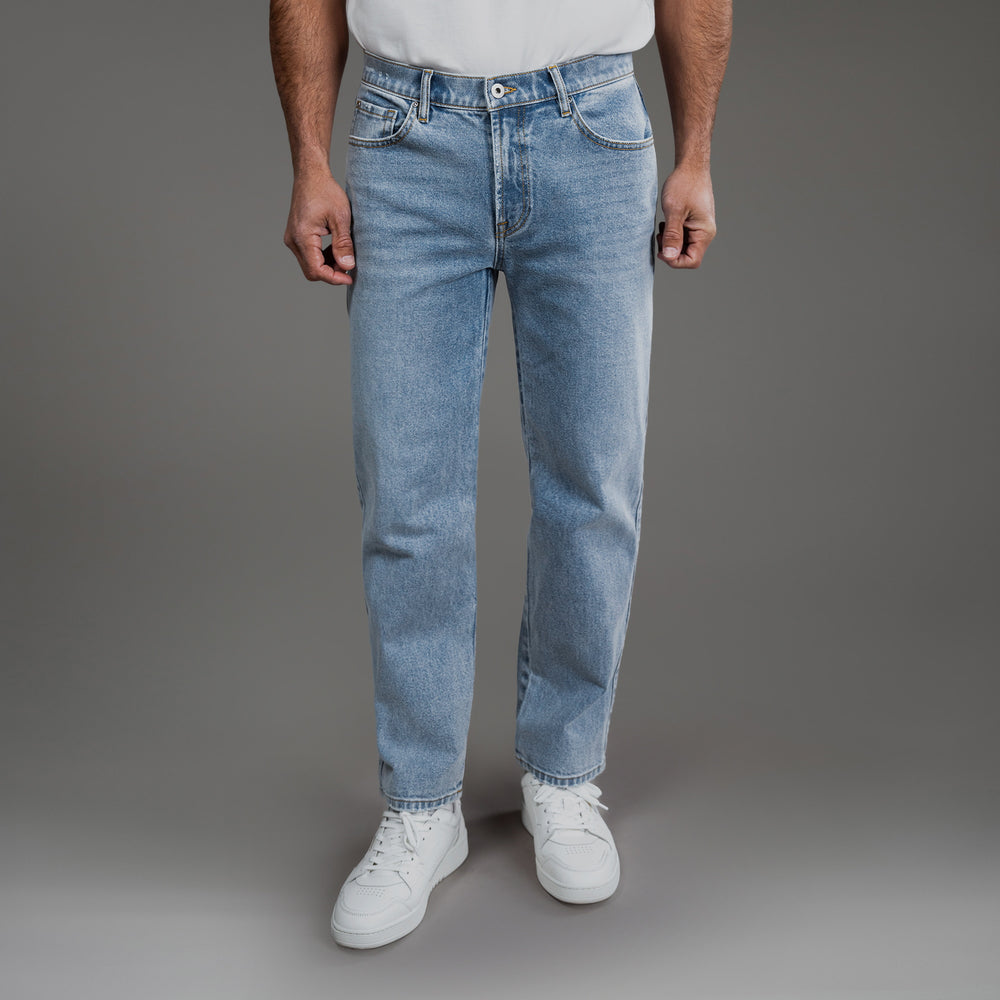 Man wearing light blue jeans and white sneakers standing against a gray background.