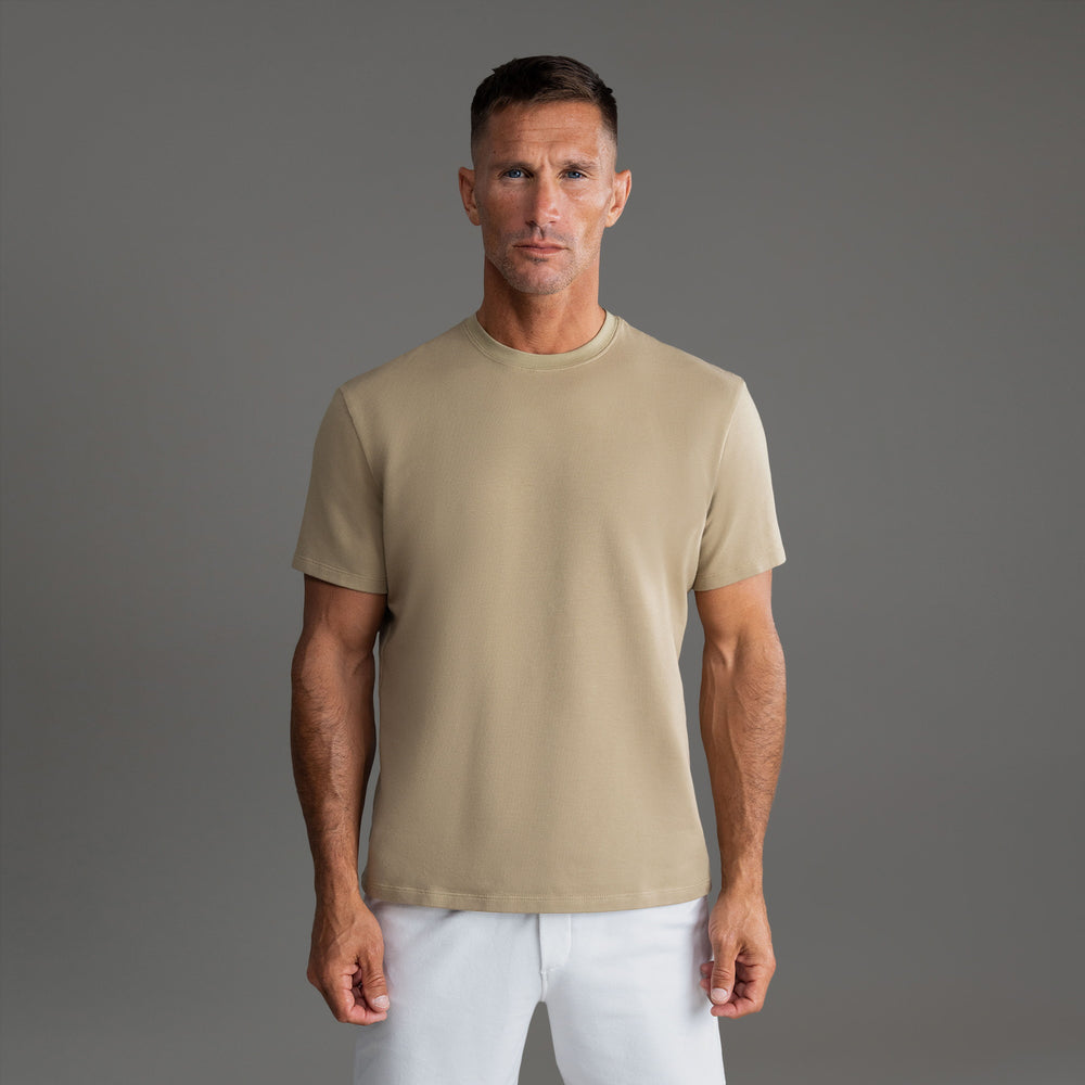 A man wearing a beige T-shirt stands against a plain gray background.