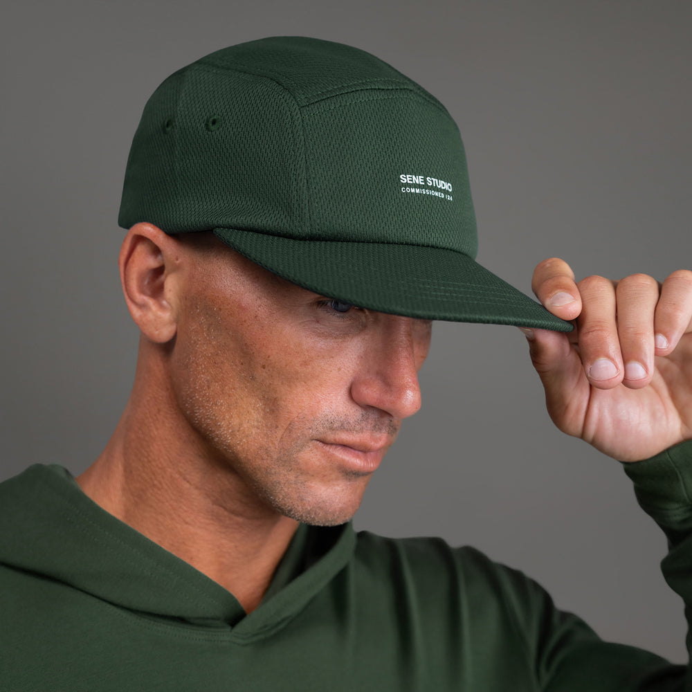 Man wearing a green cap and hoodie, holding the cap's brim.