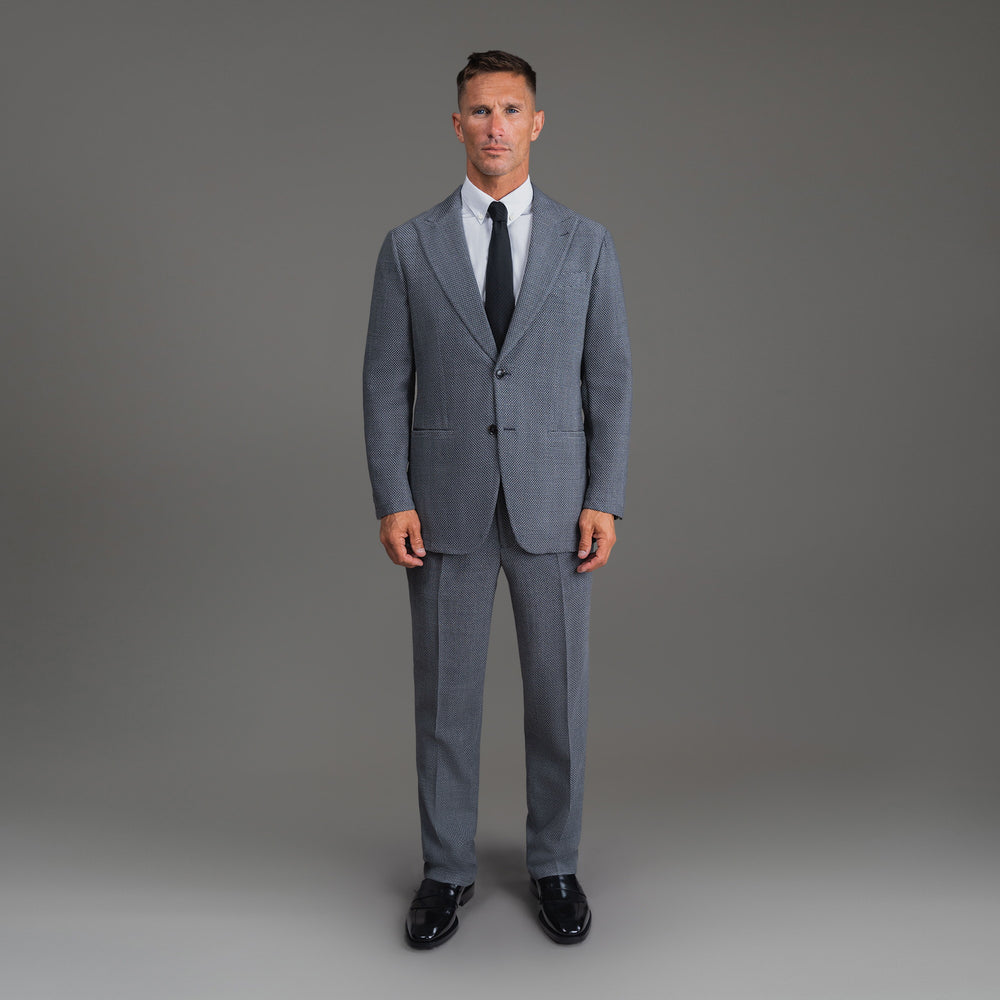 Man in grey suit, white shirt, and black tie standing against a plain background.