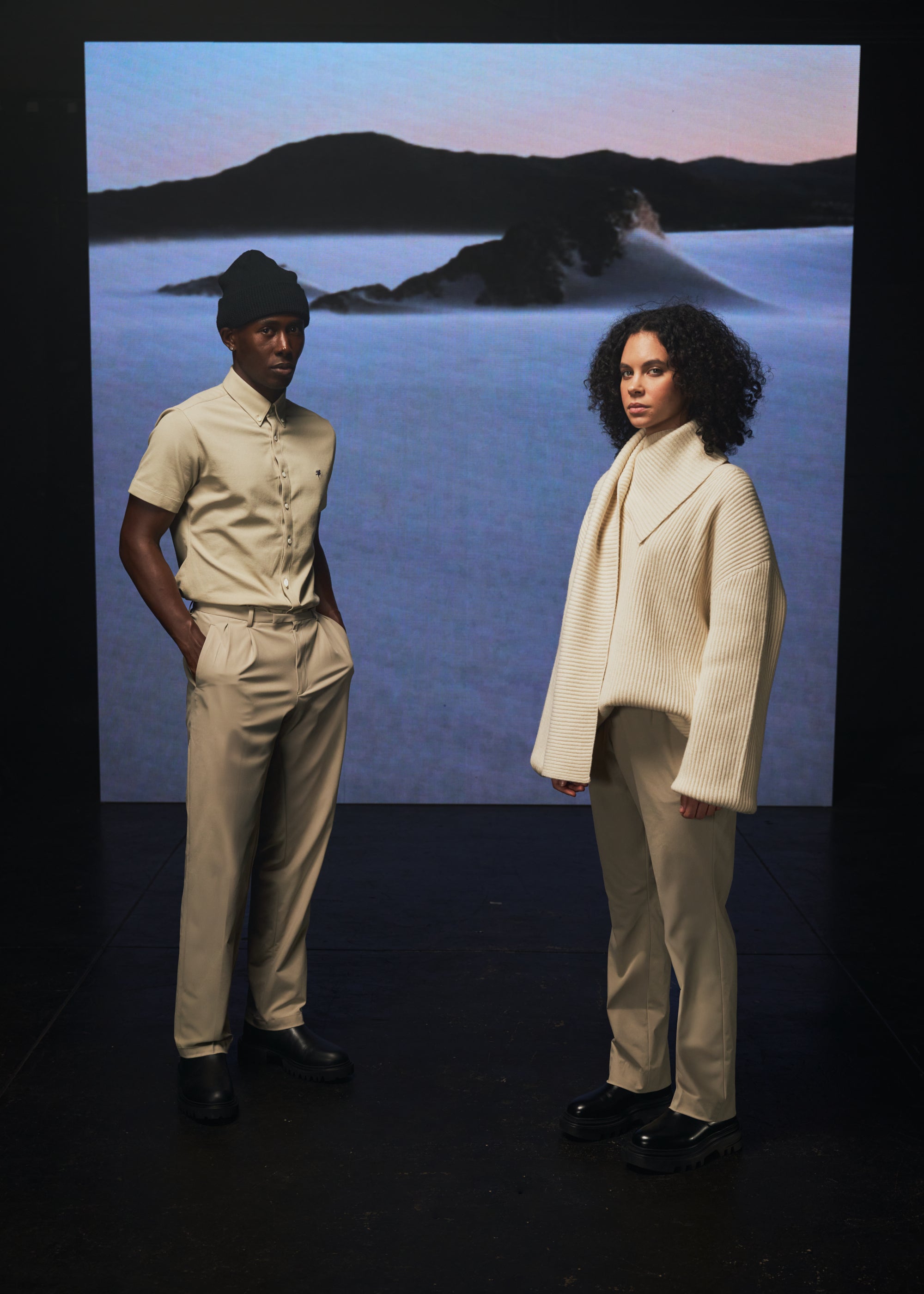 Two models in beige outfits standing against a scenic backdrop.