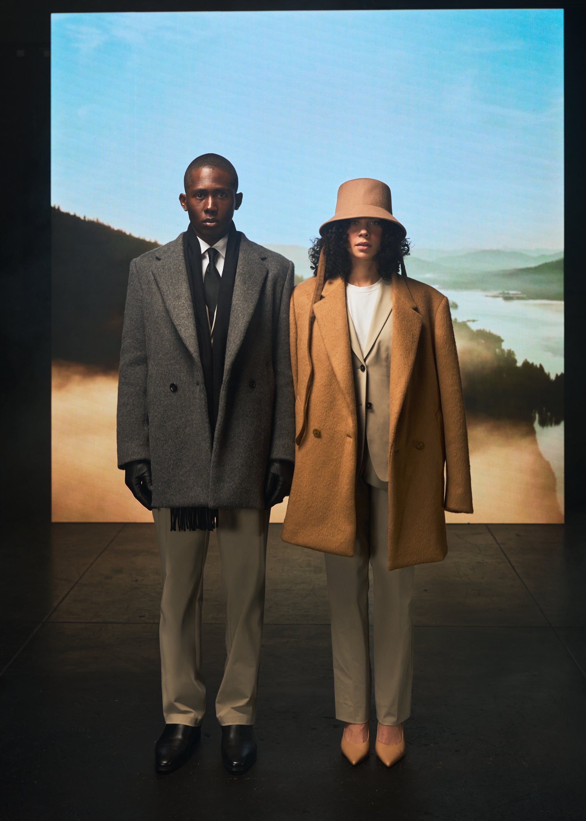 Two people in winter coats stand against a scenic backdrop.