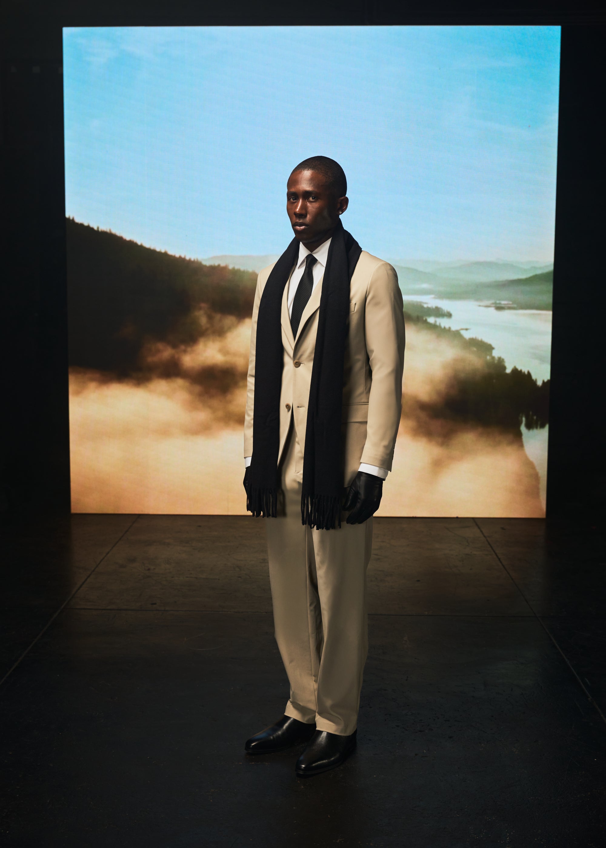 Man in beige suit and black scarf stands before misty landscape backdrop.