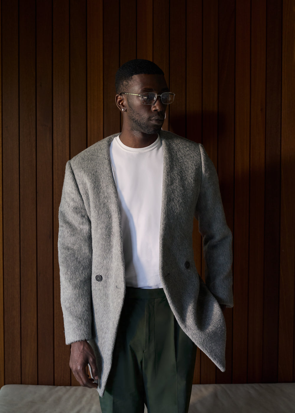 Man in a gray coat and white shirt stands against a wooden wall.