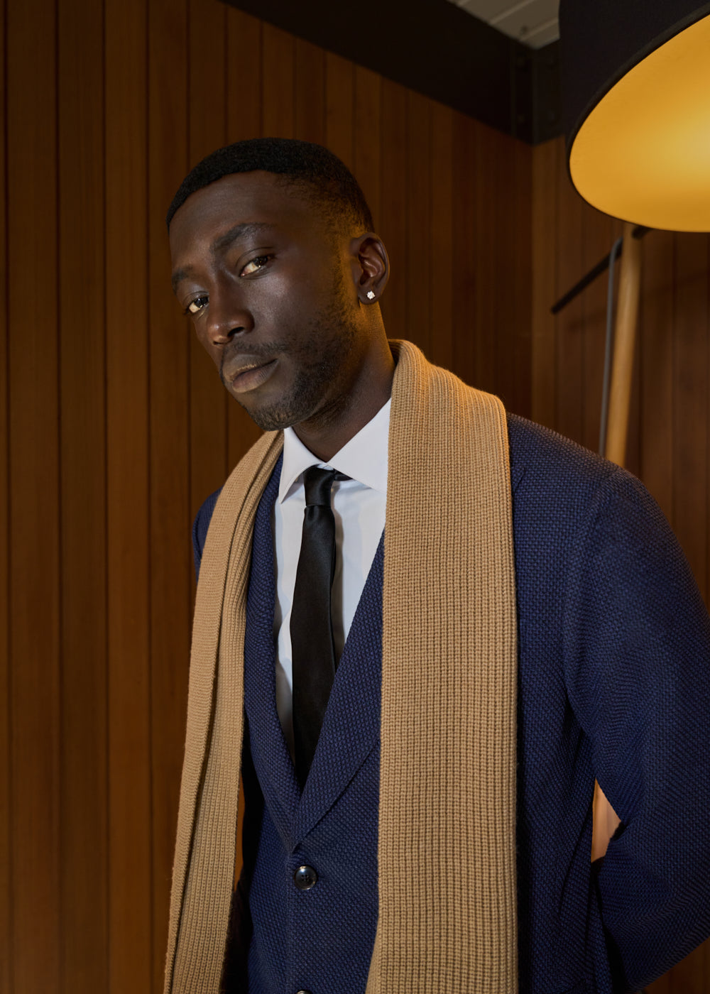 Man in blue suit, beige scarf, standing against a wooden wall with a lamp nearby.