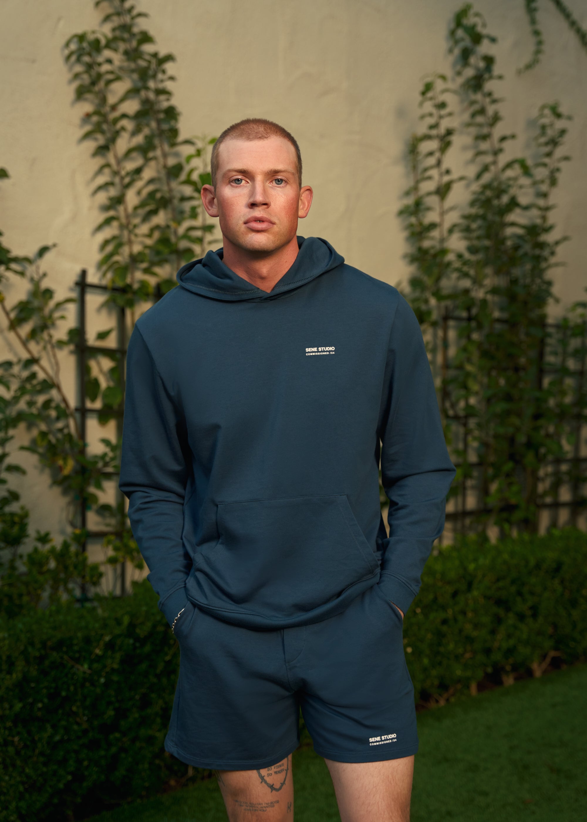 Man in blue hoodie and shorts, standing in a garden.