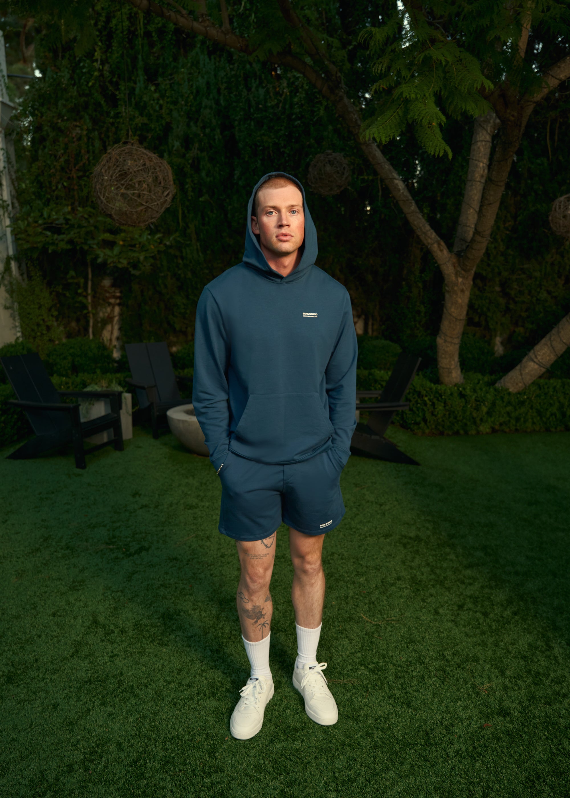 A person in a blue hoodie and shorts stands on grass with trees in the background.