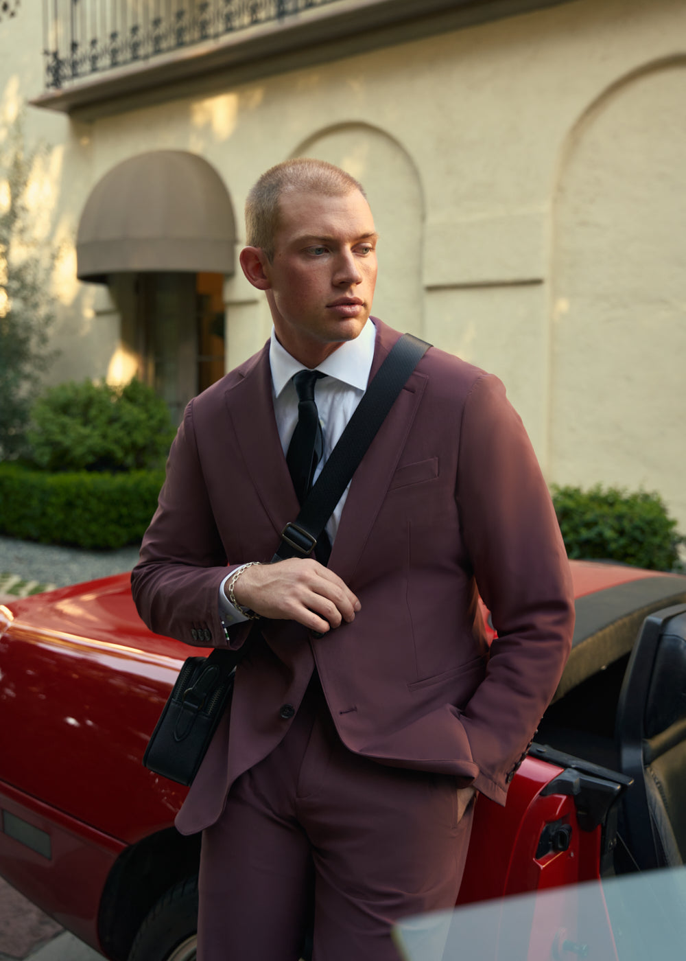 Man in maroon suit standing by a red car in front of a building.