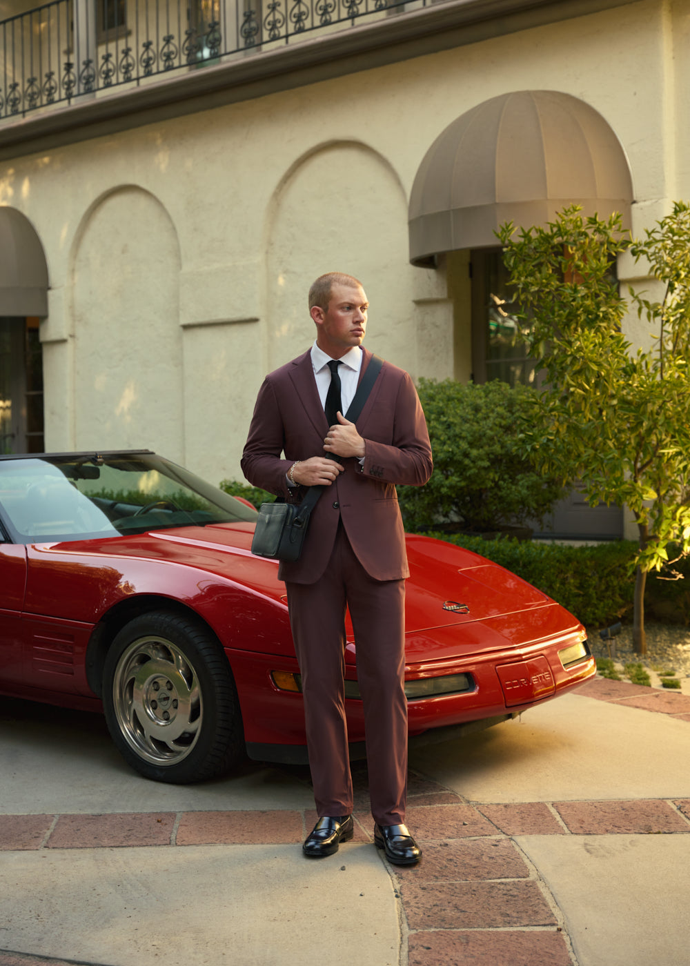 Man in a suit standing next to a red Corvette in a residential driveway.