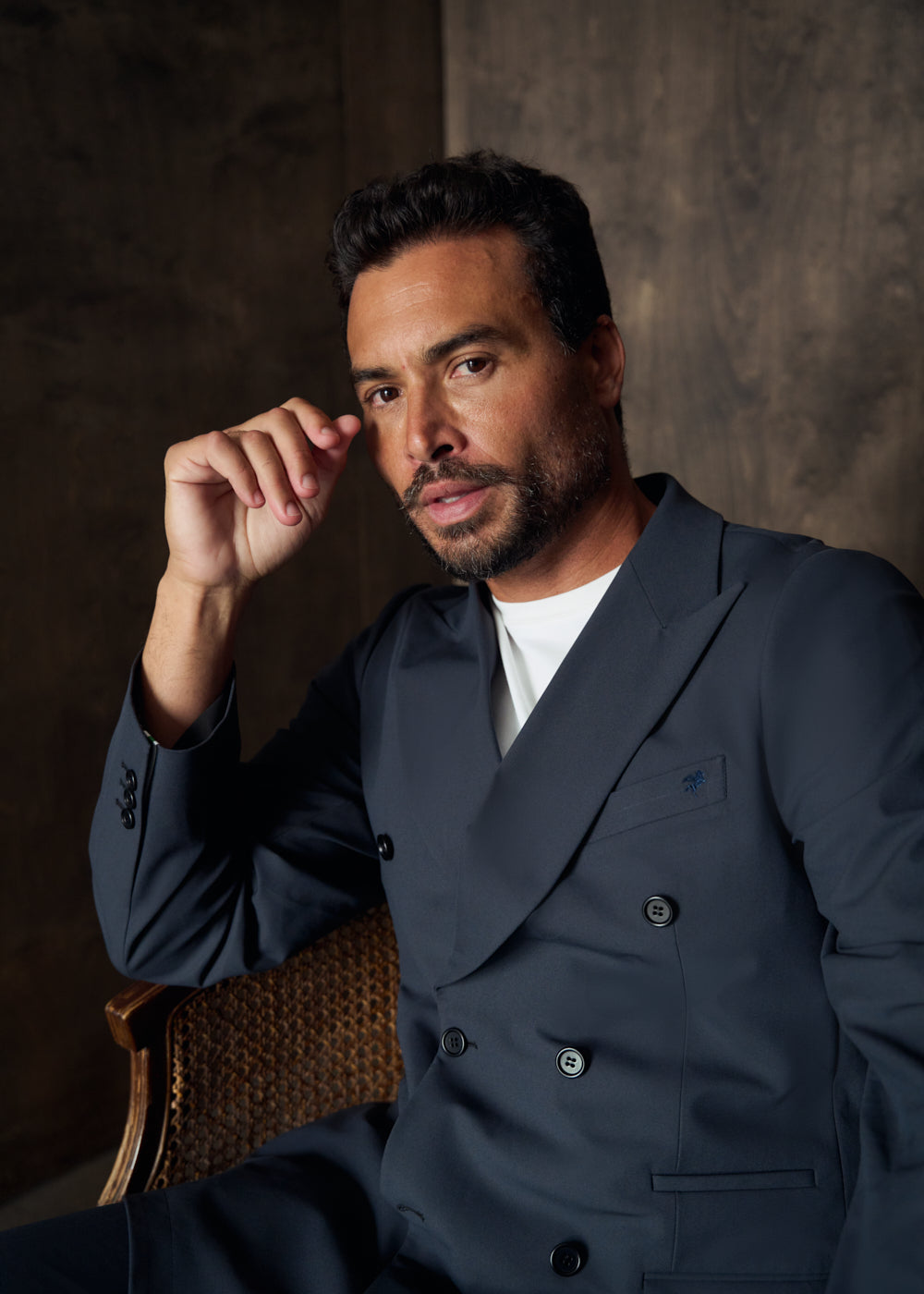 Man in a navy suit sitting on a chair, posing thoughtfully.