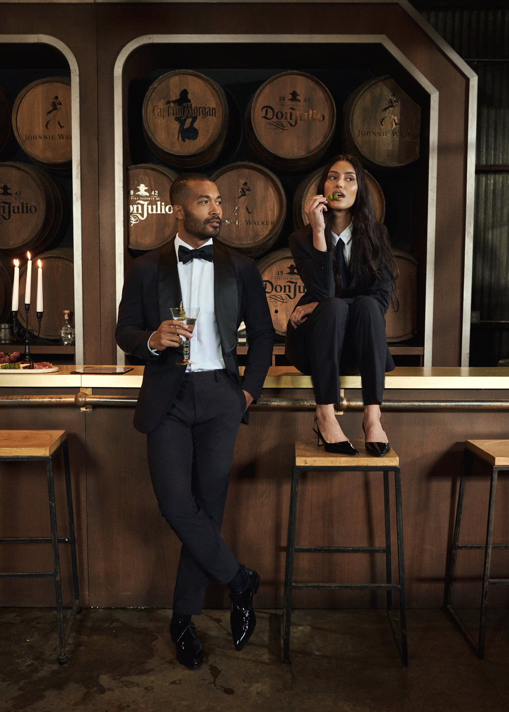 Two people in formal attire at a bar with wooden barrels in the background.