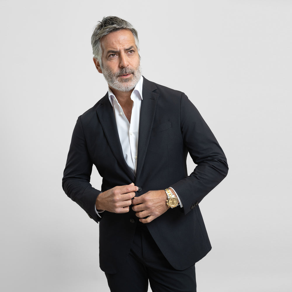 Man in a black suit adjusting his jacket against a plain background.