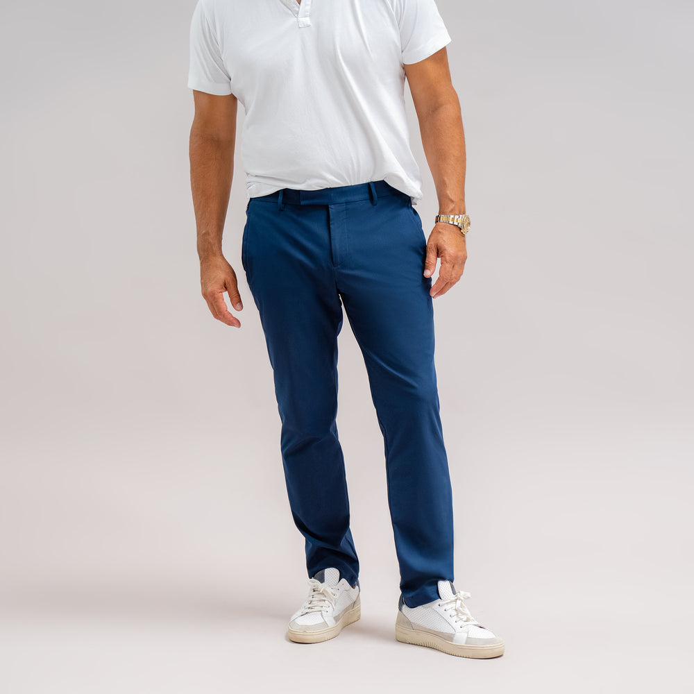 Man in white polo shirt and blue pants with white sneakers standing against a grey background.