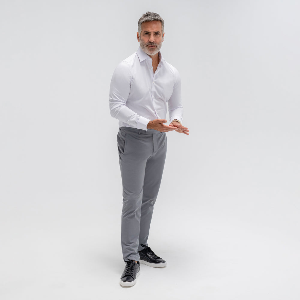 Confident man in white shirt and grey trousers standing against a white background.